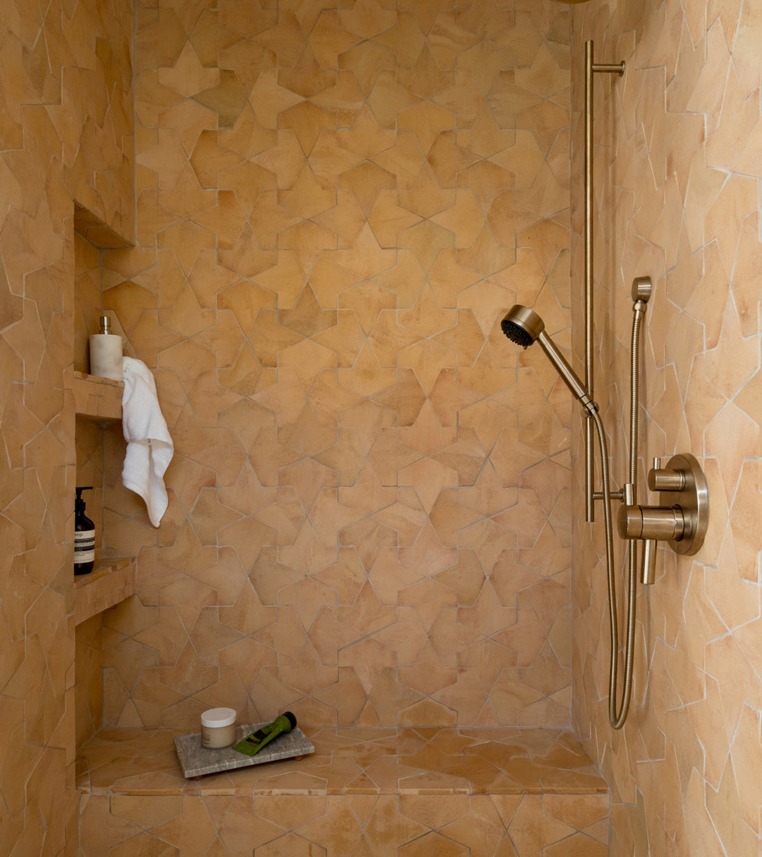 Shower stall featuring Big Alcazar + Adobe star-shaped tan wall tiles and brass fixtures, including a handheld shower head and recessed shelving.
