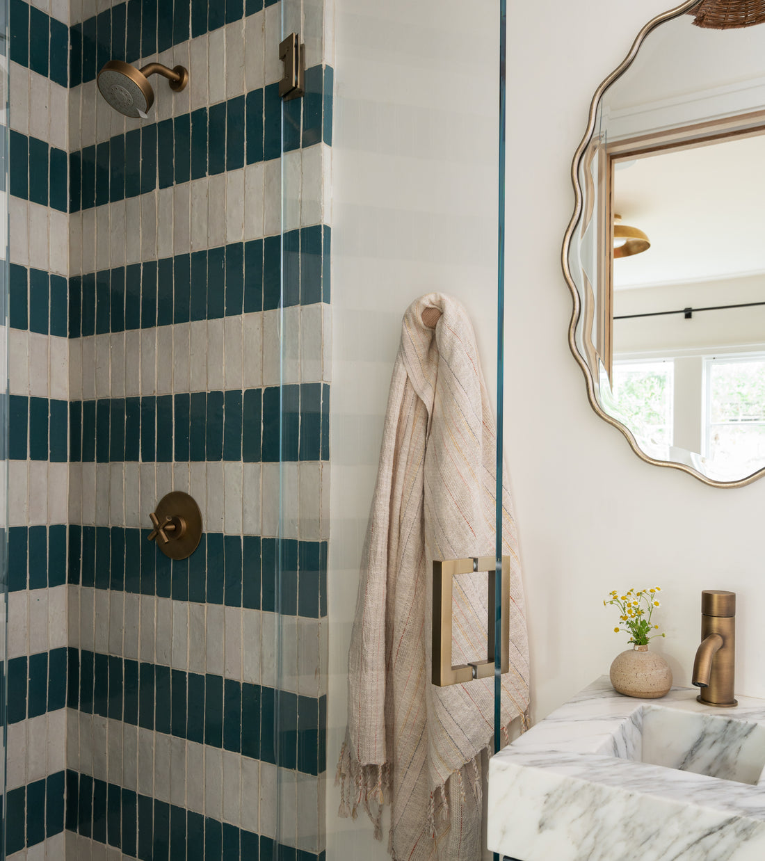 A modern bathroom featuring a shower with Zia Tile’s Aegean 2x6 blue and white zellige tiles, a brass showerhead and handle, a towel on a hook, a marble sink, a brass faucet, and a wall mirror.