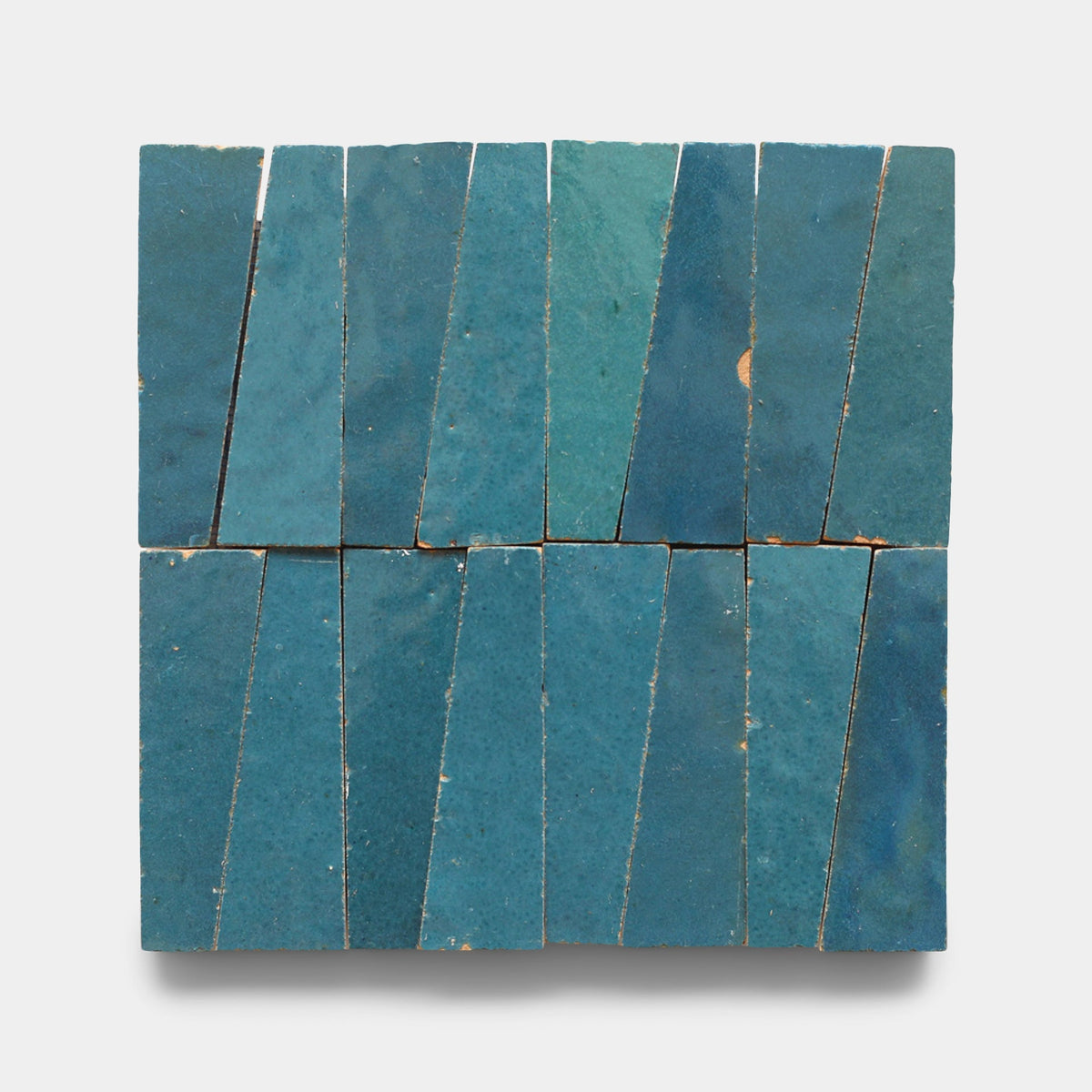 Twelve Aegean Trapezoid samples by Zia Tile are arranged in a grid. Each handmade, blue tile is angled, forming a staggered geometric pattern. The matte glaze shows slight variations and natural imperfections.