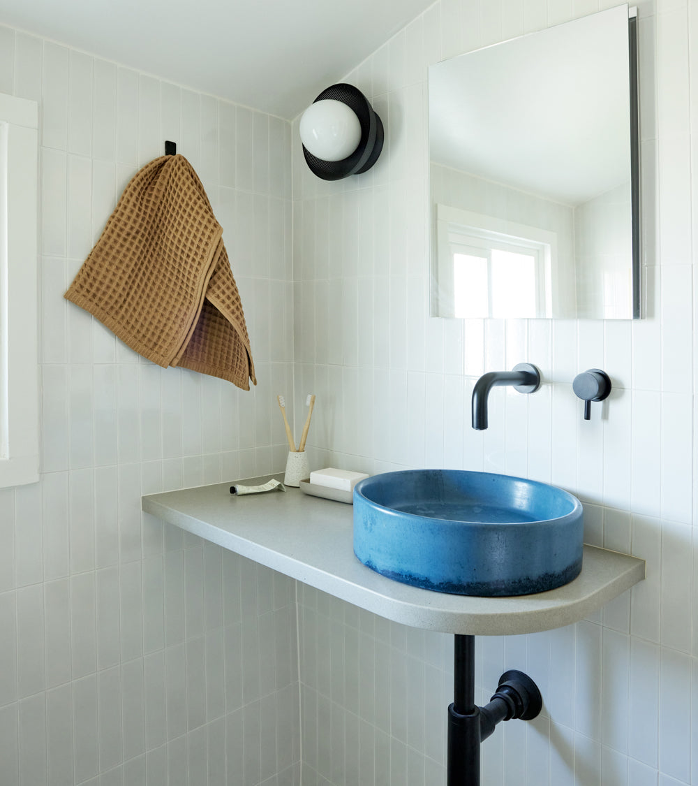 A modern bathroom features Zia Tile’s Alabaster White 2x8 glossy ceramic tile walls, a square mirror, black wall-mounted faucet, round blue sink on a gray countertop, brown textured towel on a black hook, and a round wall light above.