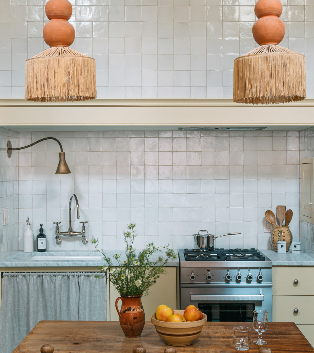 A rustic kitchen features a handmade terra cotta backsplash with Albar 4x4 Square tiles by Zia Tile, a farmhouse sink, stainless steel stove, clay pendant lights, and a wooden table displaying fruit, glassware, and a vase with greenery.