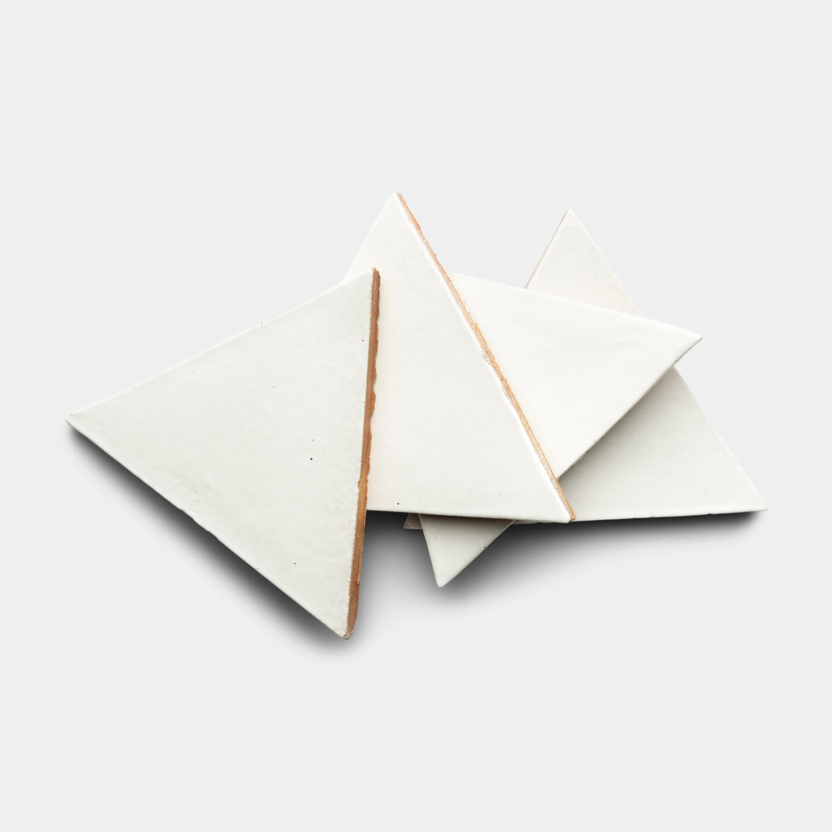 Three white Albar Triangle ceramic tiles by Zia Tile overlap on a plain light gray background, with their edges displaying a brown inner layer.