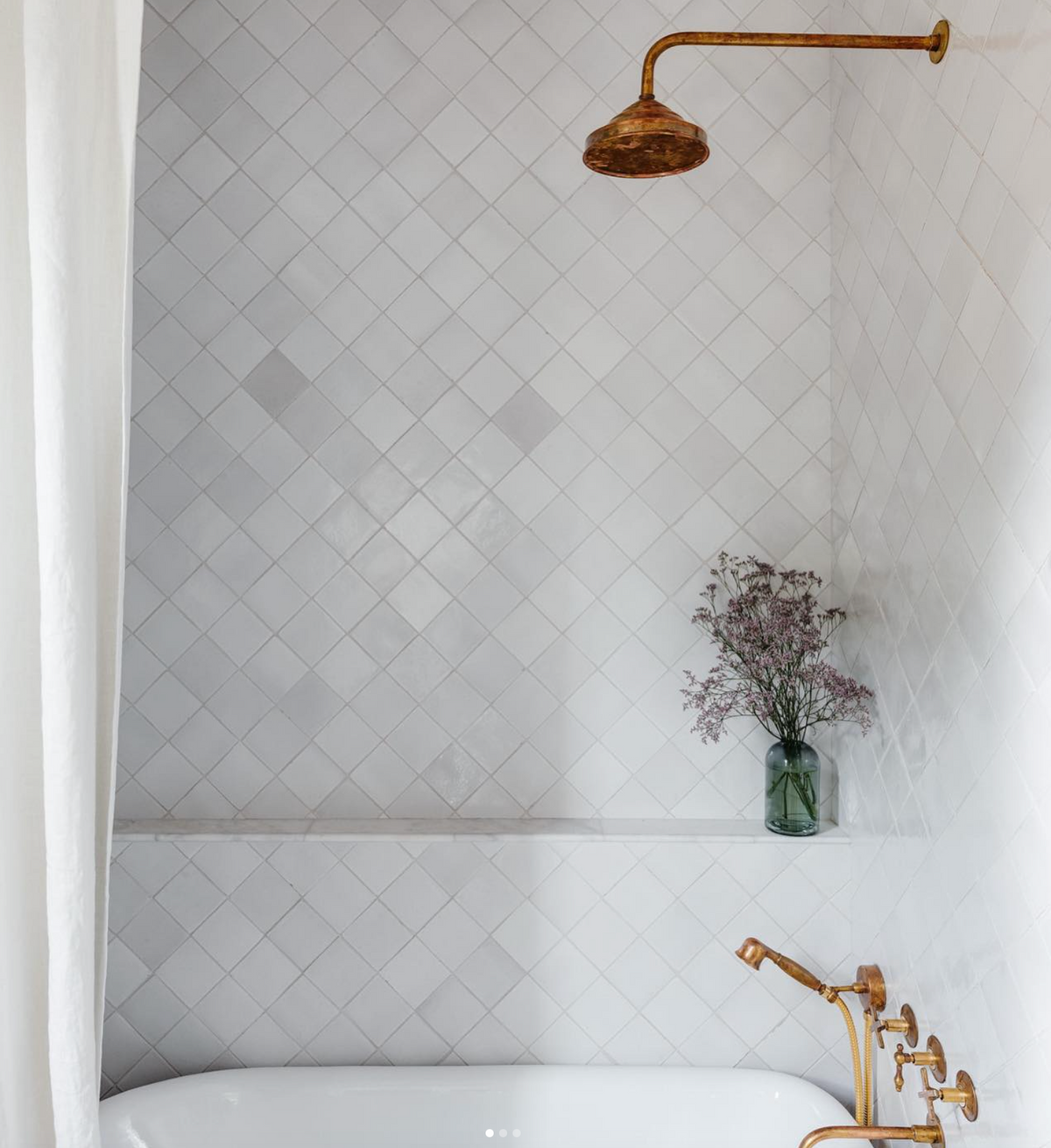 A minimalist bathroom featuring Zia Tile's Albar 4x4 Square ceramic tiles, brass fixtures, a bathtub, and a glass vase with purple flowers on a built-in shelf channels the elegance of San Miguel de Allende.