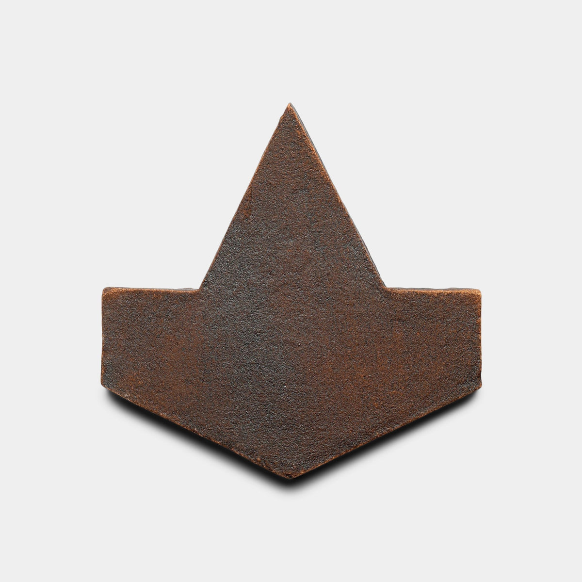 A flat, dark brown, rust-textured object shaped like a geometric arrowhead—reminiscent of a handmade tile—is shown against a white background. Product: Alcazar + Madera, Sample by Zia Tile.