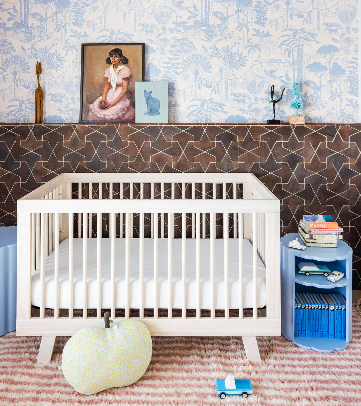 A modern nursery featuring a light wood crib, patterned rug, plush toy, and blue nightstand. A shelf displays art and decor with handmade Alcazar + Madera terra cotta tile from Zia Tile, inspired by San Miguel de Allende.