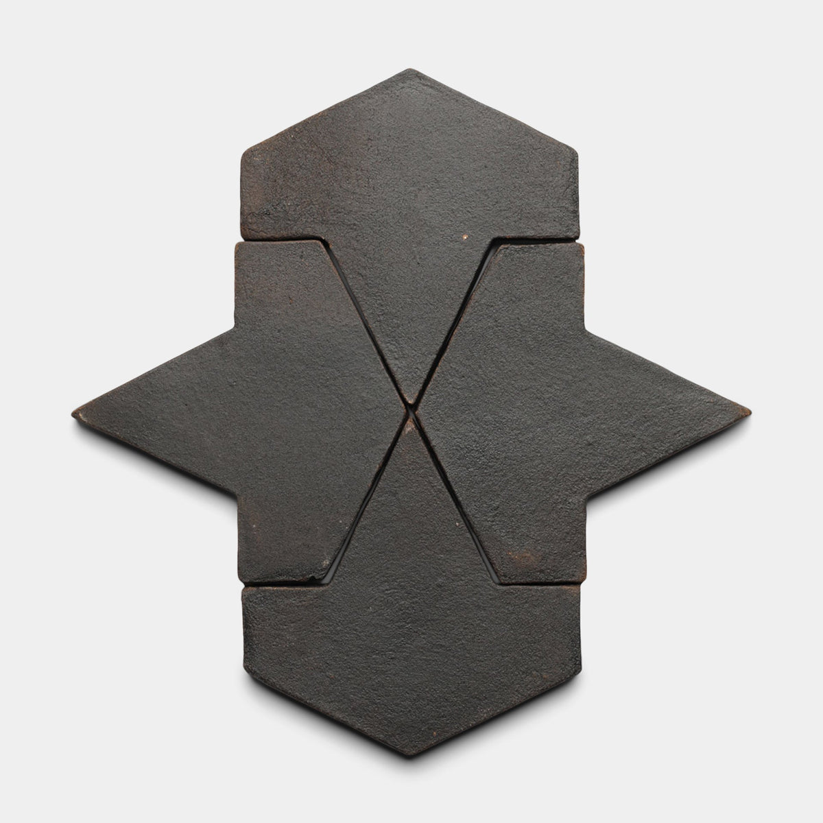 Five Alcazar + Oscura, Sample tiles by Zia Tile feature a dark, matte finish and geometric shape, arranged on a white background to form a sharp-edged, abstract star or arrow pattern with slight texture.