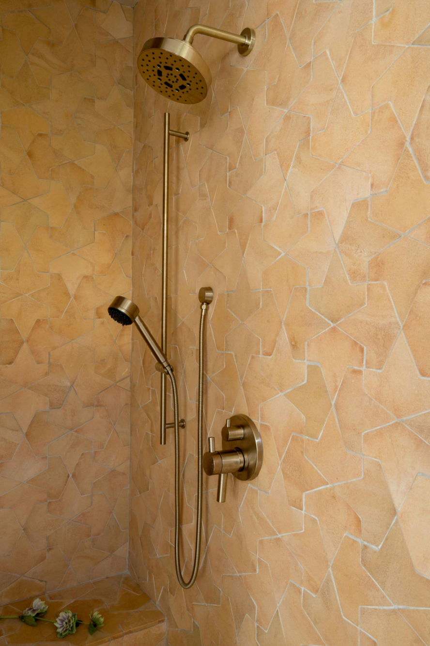 A modern shower with gold fixtures, including an overhead rain shower and a handheld showerhead, set against tan, mosaic-patterned tiles. There is a small built-in bench in the corner with a few flowers on it.
