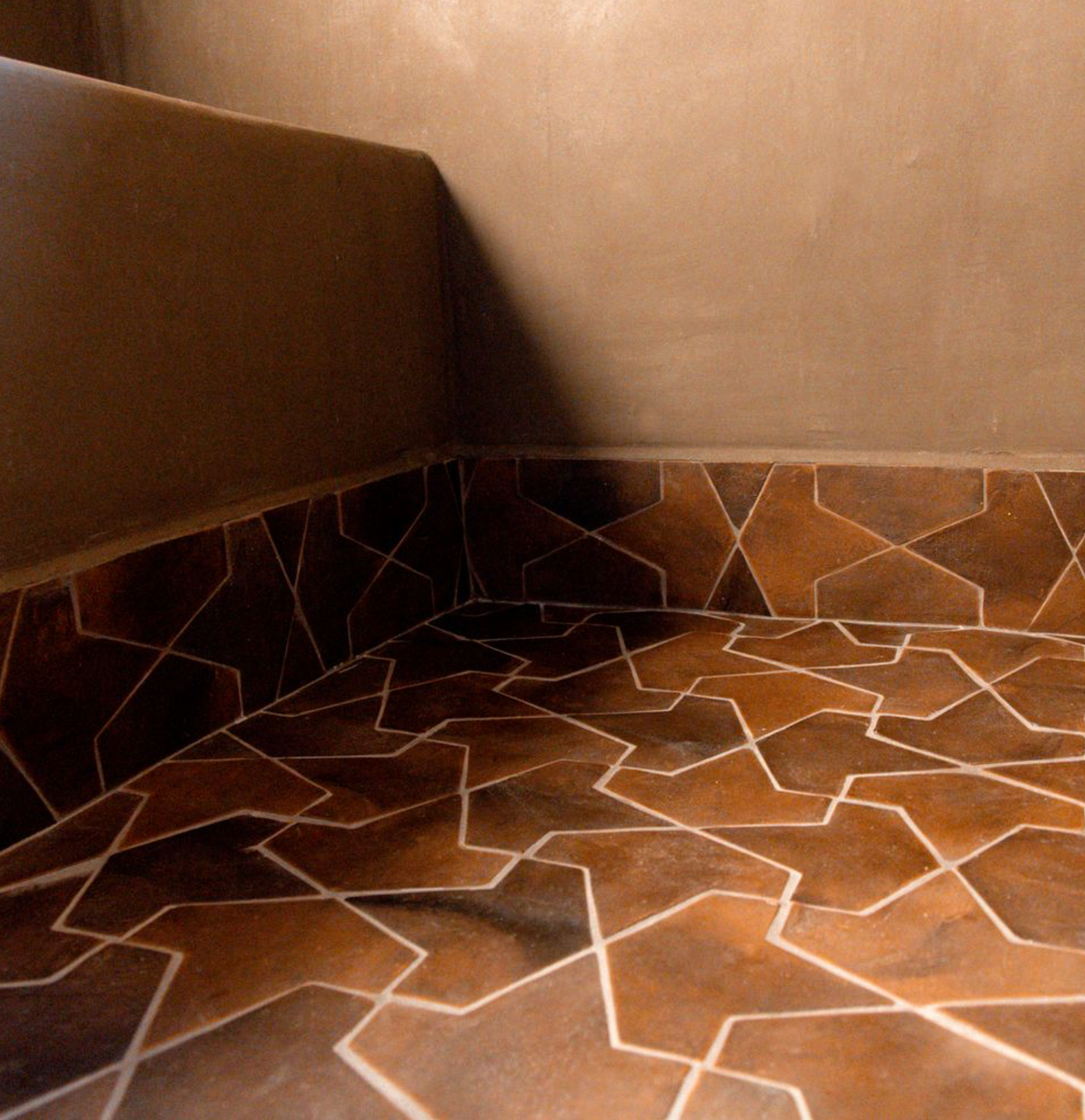 A close-up of a room’s corner showcases Zia Tile’s Alcazar + Madera sample, featuring brown geometric floor tiles with intersecting white star-like lines against smooth light brown walls, evoking Moroccan-inspired designs.