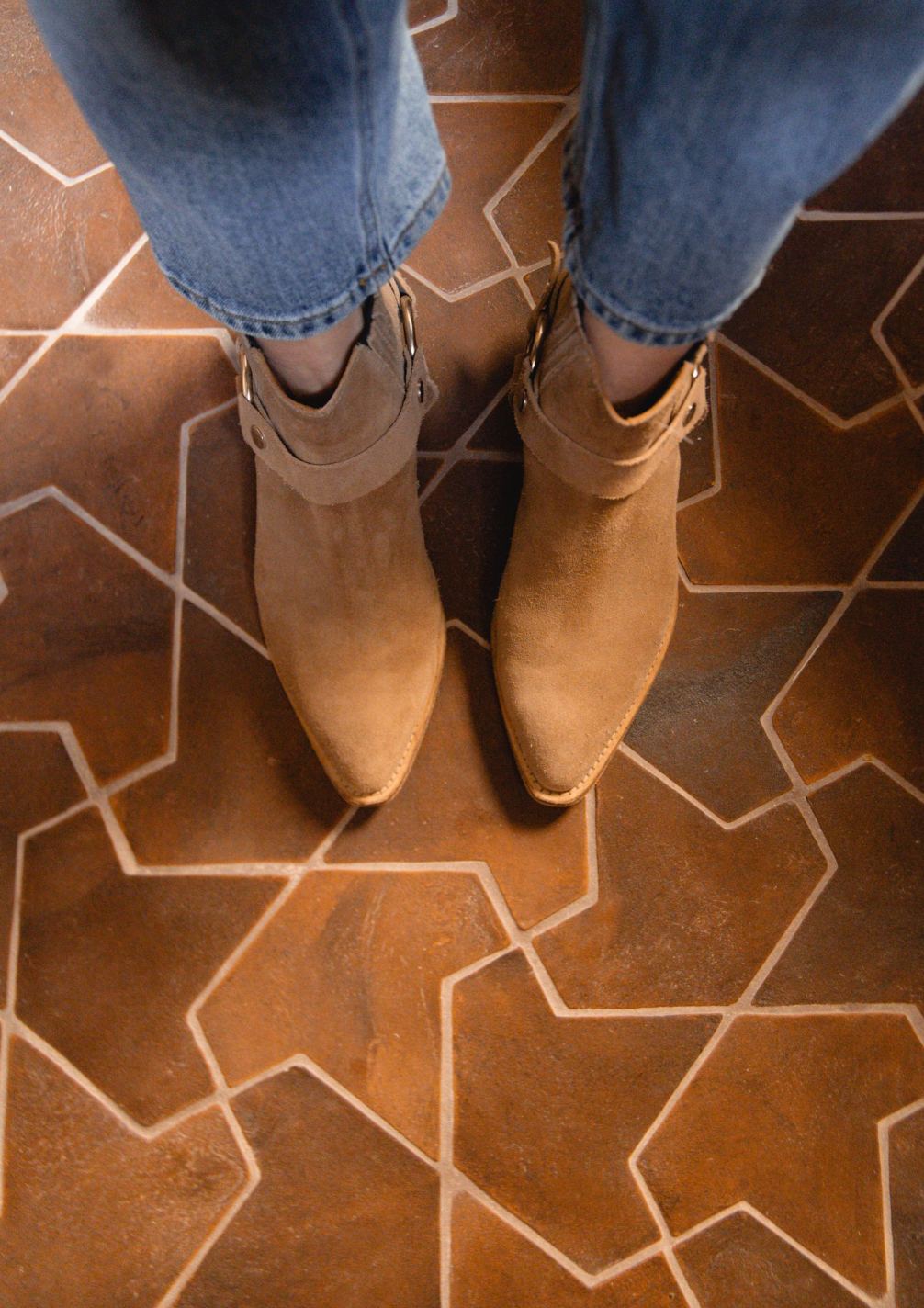 Someone wearing tan suede ankle boots and blue jeans stands on a geometric brown Moroccan tile floor featuring the Alcazar + Madera sample by Zia Tile.