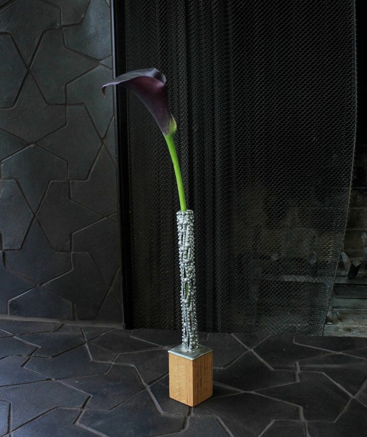 A single dark purple calla lily in a tall, textured silver vase with a wooden base is set against Zia Tile's Alcazar + Oscura sample and a black mesh screen.