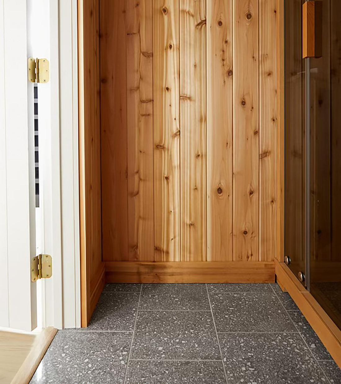 A corner with natural wood-paneled walls and a glass door sits beside a floor finished in Alexander 12x12 terrazzo cement tile from Zia Tile. Bright lighting highlights the warm wood tones and modern aesthetic.