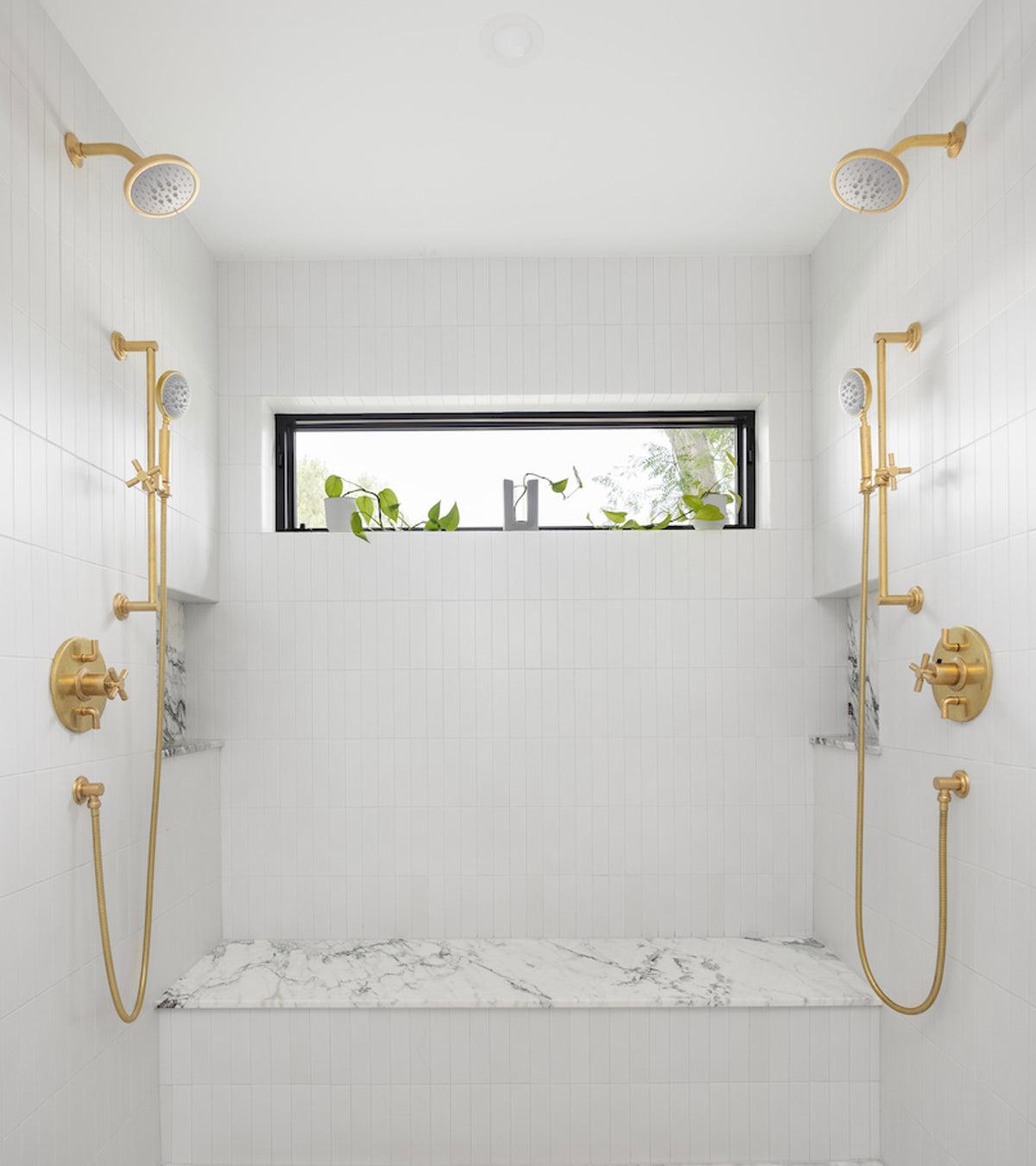 Modern double shower featuring Zia Tile’s Alpha White 2x8 ceramic tiles, gold fixtures, a marble bench, and a narrow horizontal window above showcasing greenery.