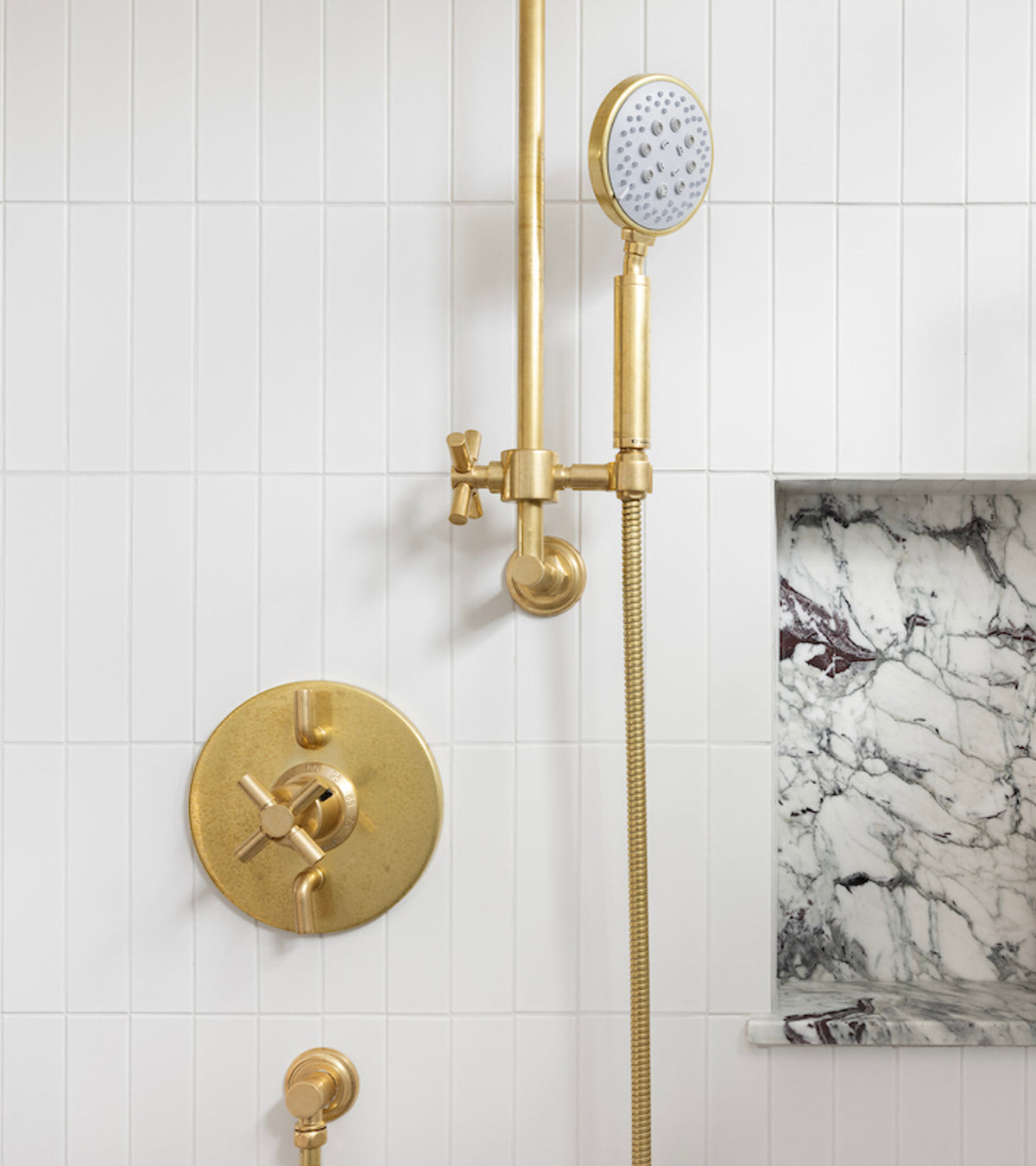 A modern shower features Zia Tile’s Alpha White 2x8 tiles with gold fixtures, including a round handheld showerhead and control knobs. On the right, a recessed shelf displays a white and gray marble background.