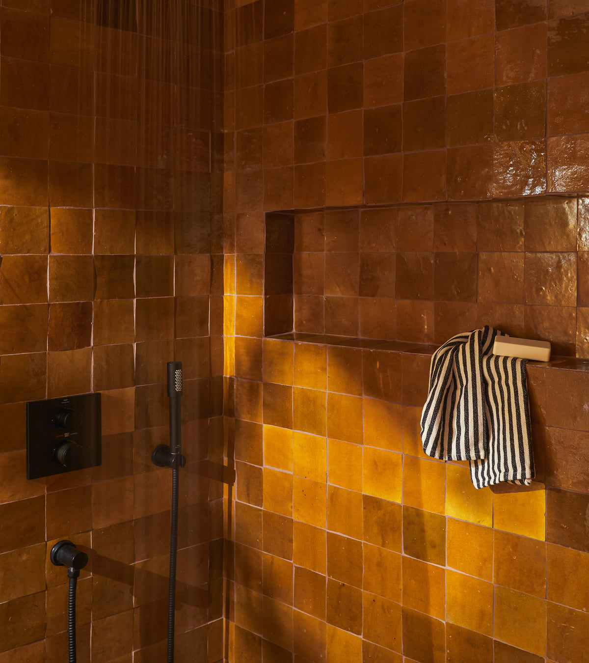 A shower features vendor-unknown’s Amber 4x4 tiles, a black shower fixture, a built-in niche, and a black-and-white striped towel on a wooden brush. Warm lighting accentuates the tile’s rich texture.