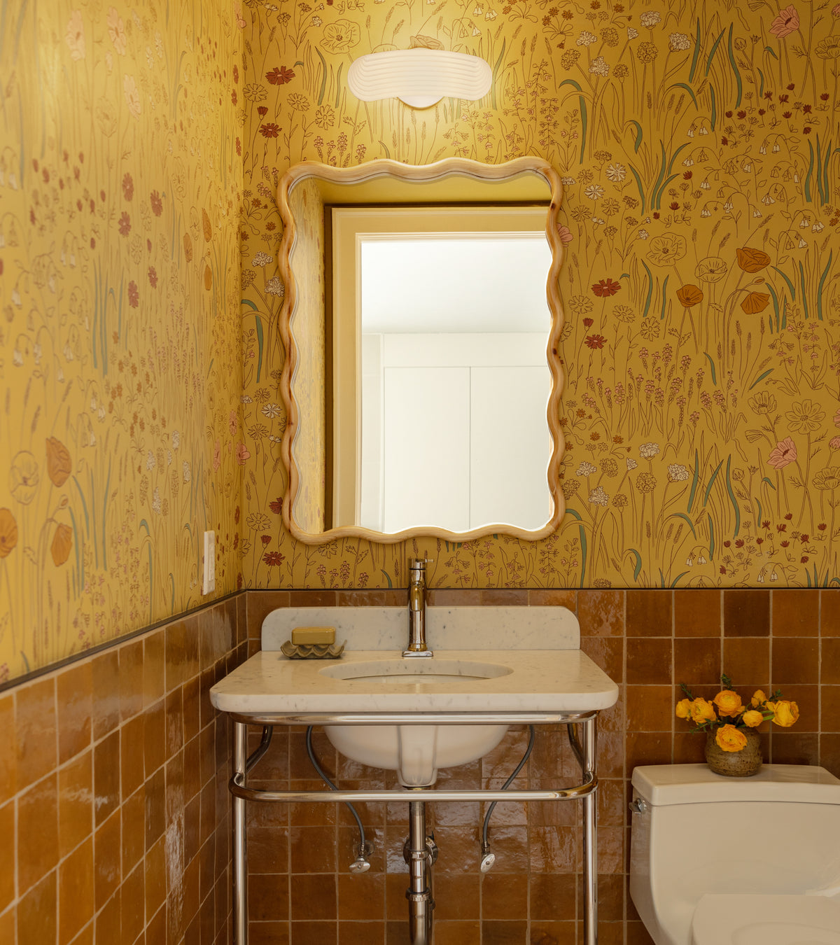 A small bathroom with yellow floral wallpaper, a white sink with metal legs, a scalloped mirror, a modern wall light above, vendor-unknown Amber 4x4 handmade mosaic tiles on the lower walls, and a toilet with yellow flowers.
