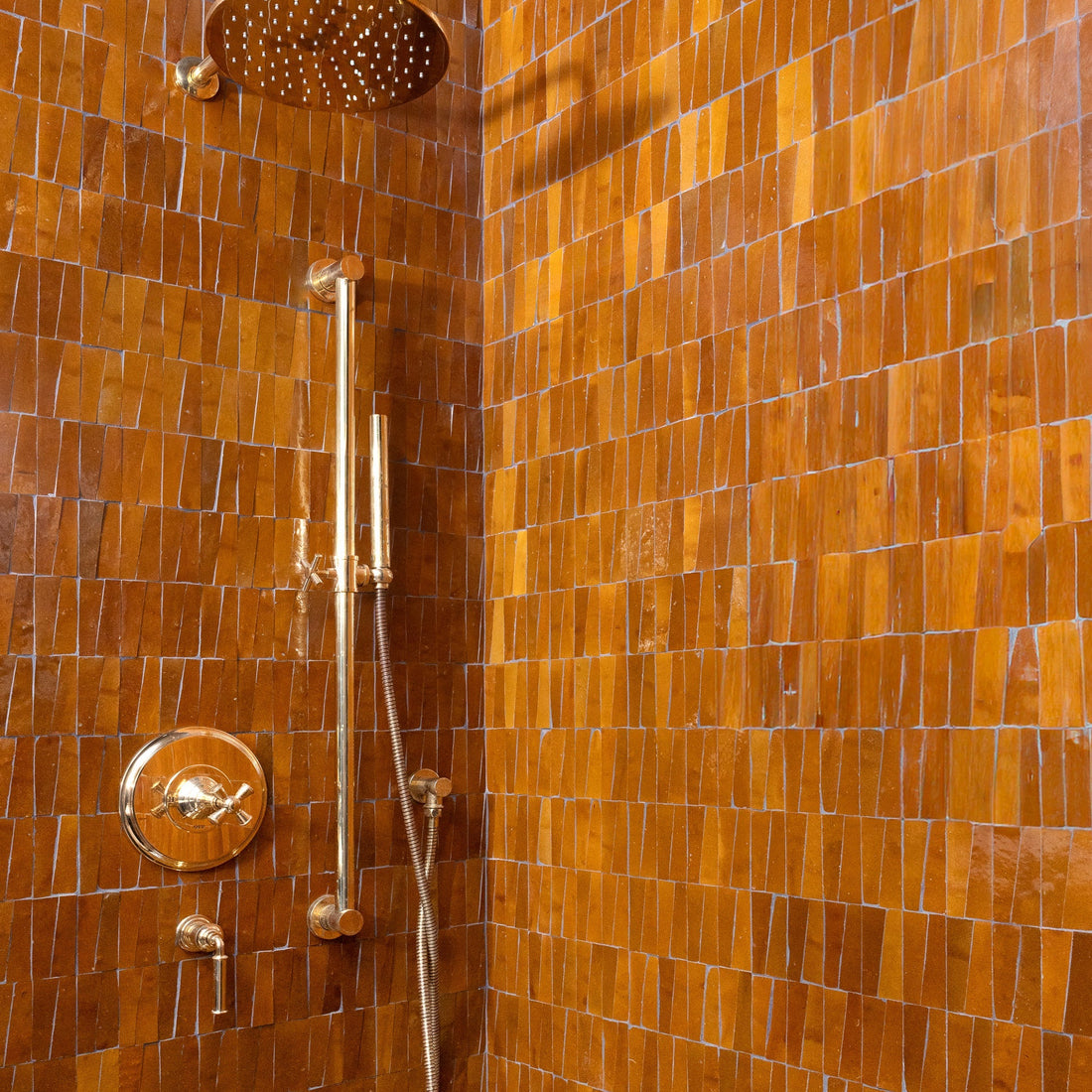 Shower features gold fixtures with a rain showerhead and hand shower, set against walls clad in Zia Tile's Amber Trapezoid tiles, shiny rectangular brown tiles arranged vertically.