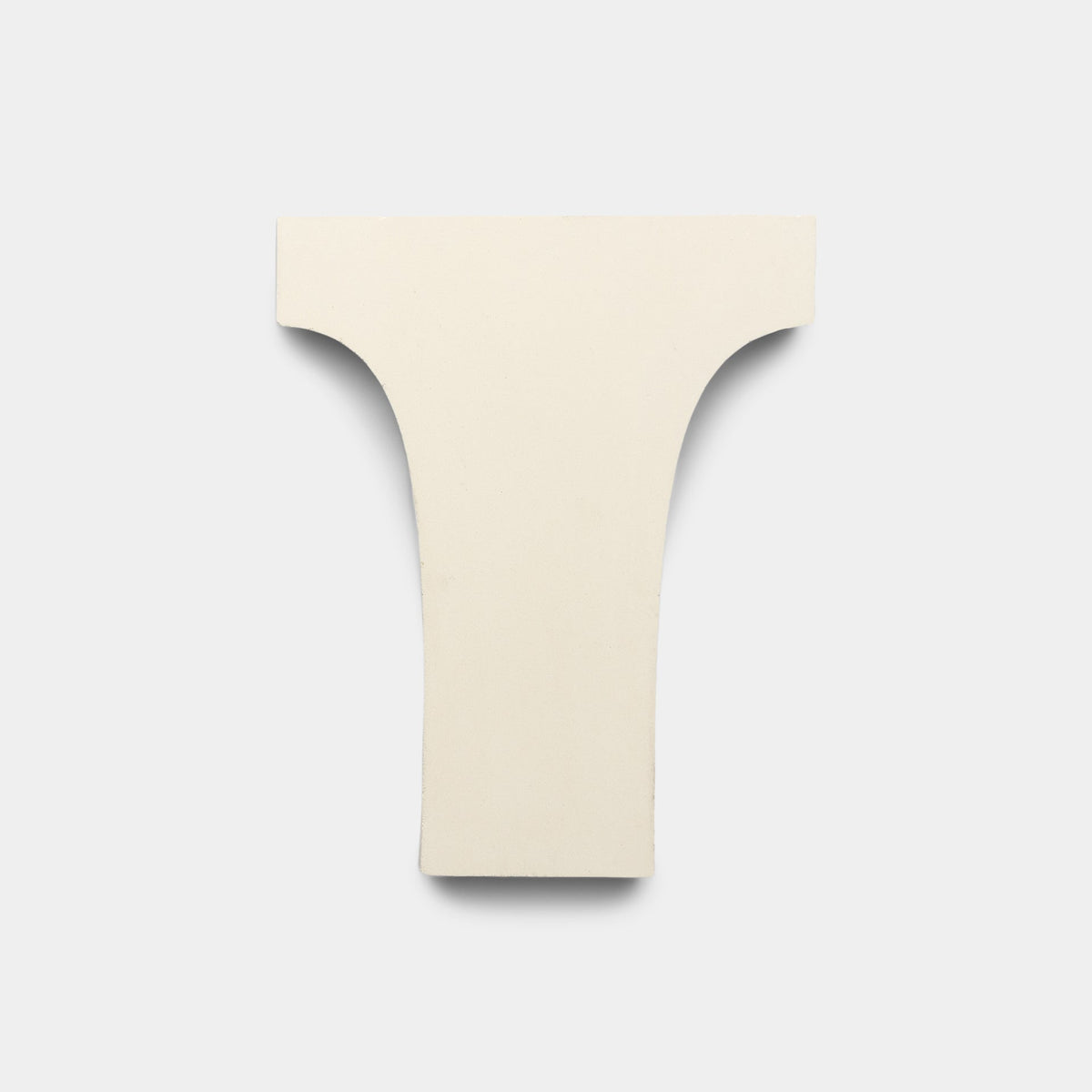 A cream-colored, T-shaped handmade tile, Arco Bone + Black + Mustard by Zia Tile, with smooth edges rests on a plain light gray background—an elegant ceramic form showcasing Zia Tile craftsmanship.