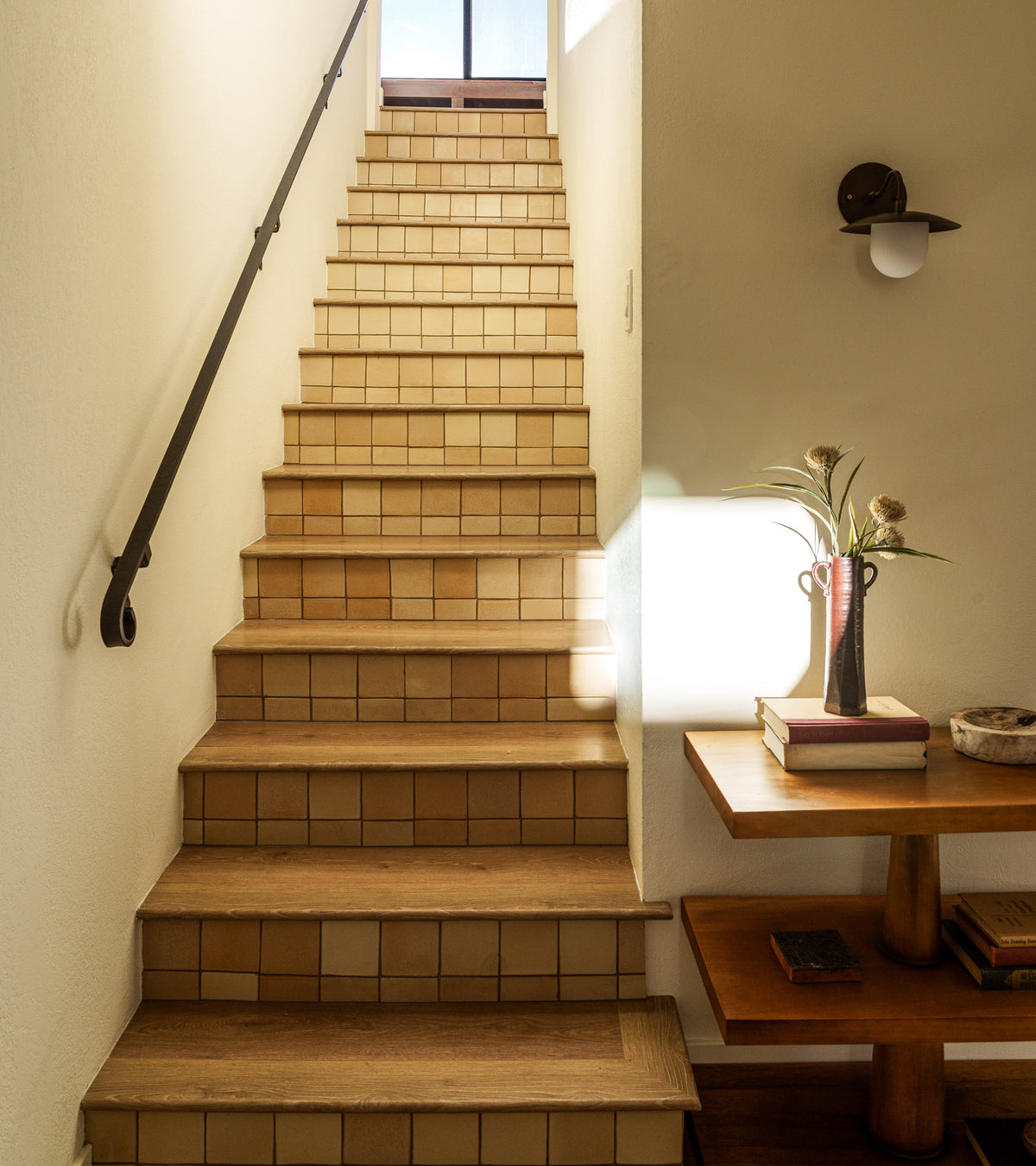A staircase with wooden steps and handmade Arroyo 4x4 Square tiles by Zia Tile leads to a sunlit window. On the right, a wooden table displays flowers, books, and a bowl, while a black handrail and wall sconce line the left wall.