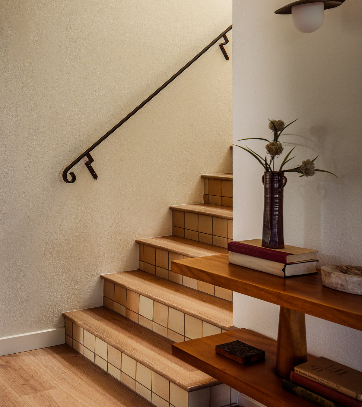 A wooden staircase with handmade Arroyo 4x4 Square tiles by Zia Tile on the risers curves upward beside a black handrail. In the foreground, a wooden shelf holds books and a brown vase with dried flowers.