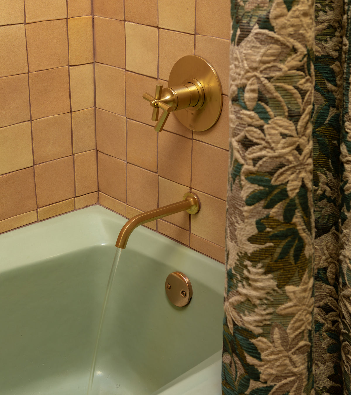 A bathtub with pale green enamel fills beneath a gold faucet. The wall showcases Arroyo 4x4 Square tiles by Zia Tile, while a floral-patterned shower curtain is partly visible on the right.