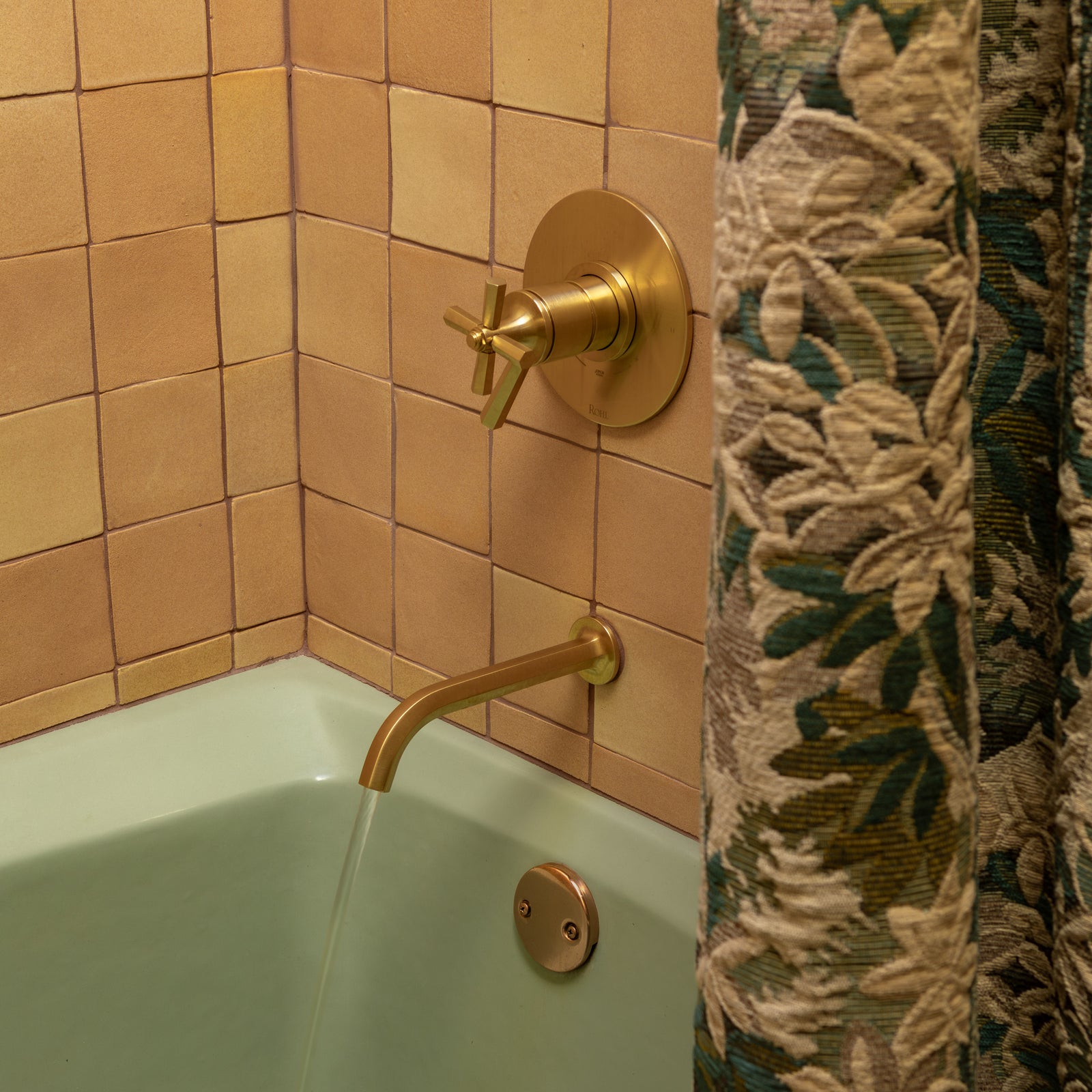 A bathtub with light green enamel, gold-colored faucet, and handle. Water flows from the spout. The wall is covered in beige square tiles, and a floral-patterned shower curtain partially covers the right side.