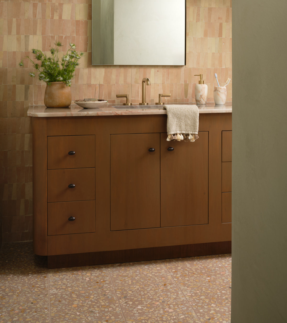 A bathroom featuring a wooden vanity, gold faucet, marble countertop, beige hand towel, potted plant, and containers. The wall is finished with Artemis 12x12 terrazzo tiles by Zia Tile and the floor has tan tiles.