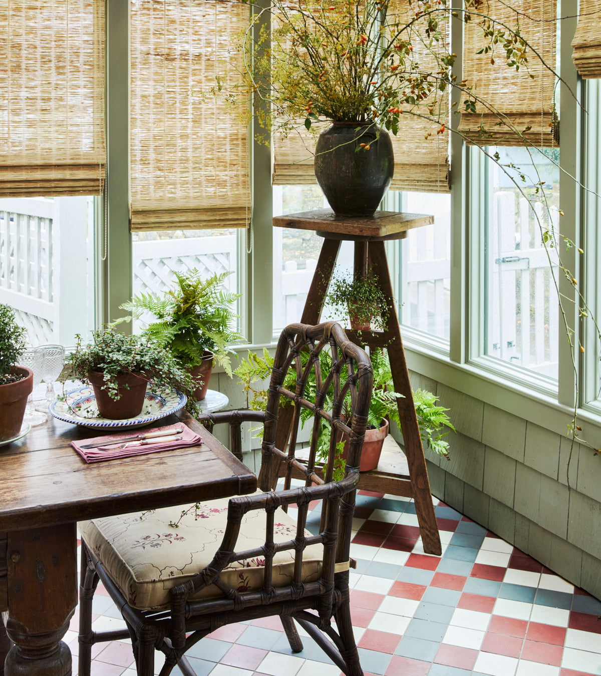 A sunlit room with large windows, bamboo blinds, a wicker chair with a floral cushion, a wooden table, potted plants, and a checkered floor featuring Zia Tile’s Ash 4x4 cement tile in red, white, and blue.