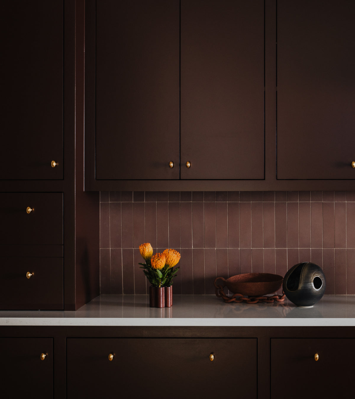 A dark brown kitchen features gold cabinet knobs, a white countertop, orange flowers, and a black and gold vase set against an Aubergine 2x8 subway tile backsplash by Zia Tile.