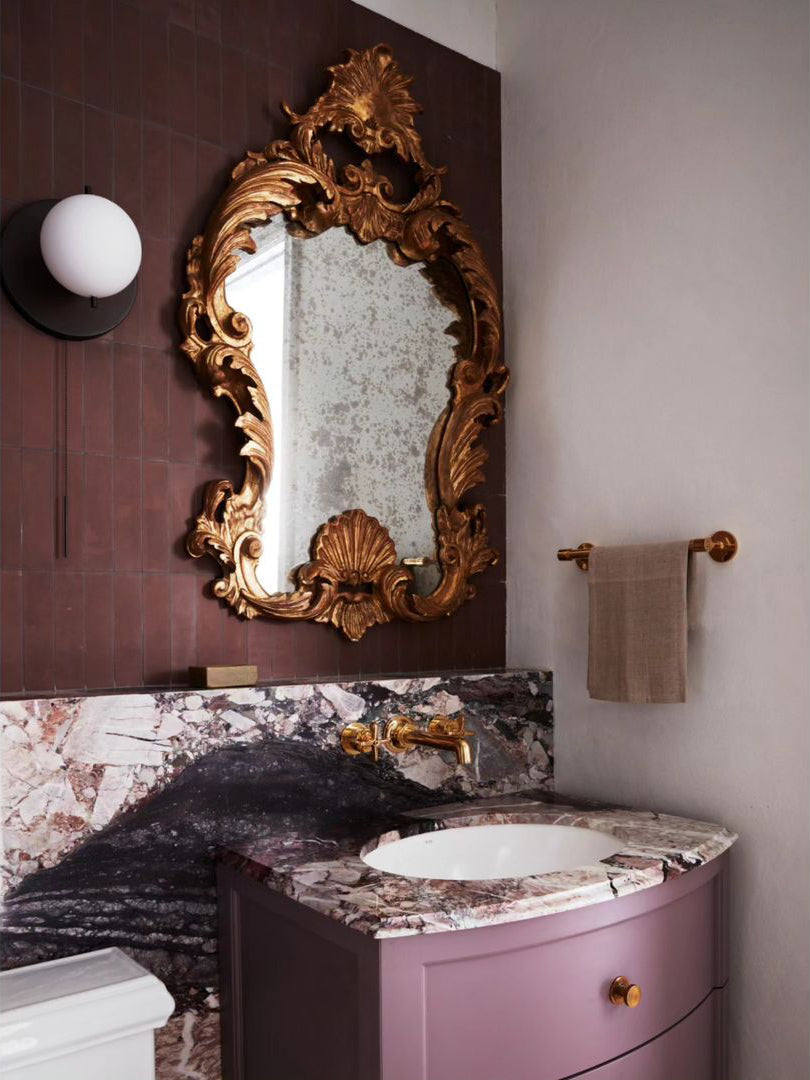 A bathroom features a marble sink and countertop, gold ornate mirror, wall-mounted faucet, round wall light, brown towel on a rack, set against mauve and off-white walls with Zia Tile's Aubergine 2x8 handmade tile accents.