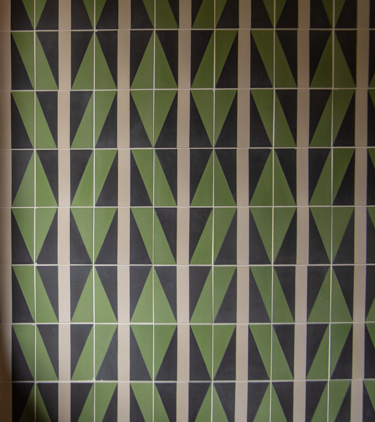 A wall adorned with Zia Tile's Aviator Olivine 4x4 tiles features a geometric arrangement of green, black, and beige triangles for a symmetrical and eye-catching design.