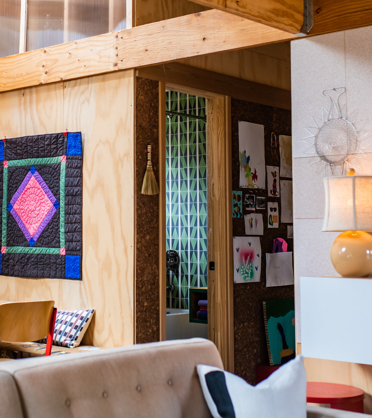 A cozy, modern living space with a quilt on a wooden wall, art and a sofa nearby, and a lamp on the side table. The doorway opens to a room featuring Aviator Olivine 4x4 tiles by Zia Tile as patterned accents against green geometric wallpaper.