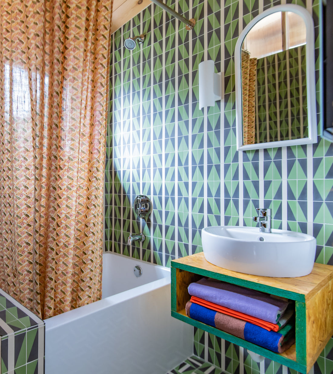 A vibrant bathroom featuring Aviator Olivine 4x4 green geometric tiles by Zia Tile, a white tub with a patterned curtain, round mirror, modern white sink on a wooden shelf, and neatly folded towels beneath.