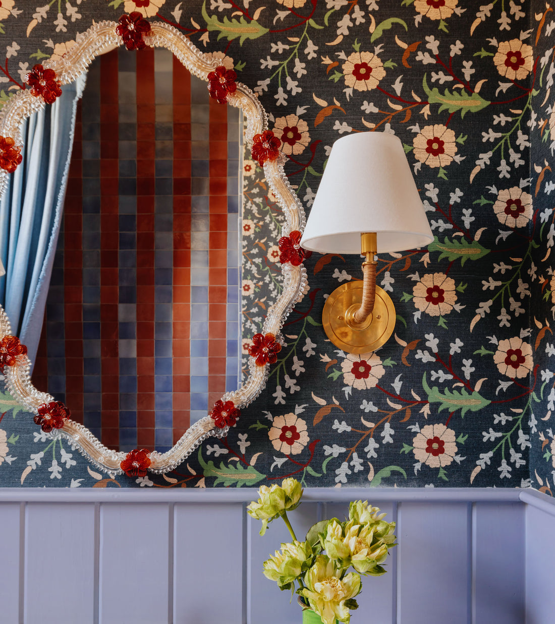 An ornate mirror with red flower accents hangs on dark floral wallpaper near a brass-white sconce. Green flowers sit on lavender wainscot, while Zia Tile’s Azul 4x4 Square tiles are reflected in the mirror.