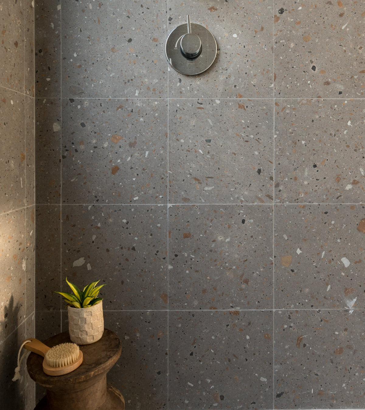 A shower with gray terrazzo tiles, a round silver showerhead, a wooden stool holding a potted plant, and a wooden bath brush resting beside the plant.