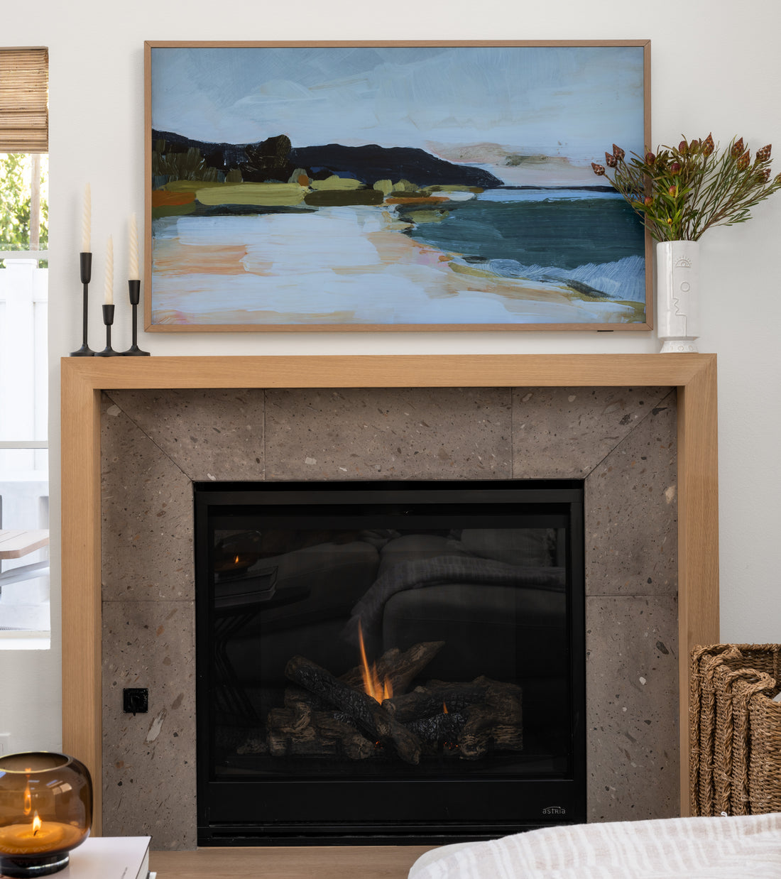 A cozy living space features a modern fireplace with a Zia Tile Badlands 12x24 gray stone tile surround and wooden mantel, topped with a coastal painting, candles, and flowers. Nearby sit a wicker basket and glass container.