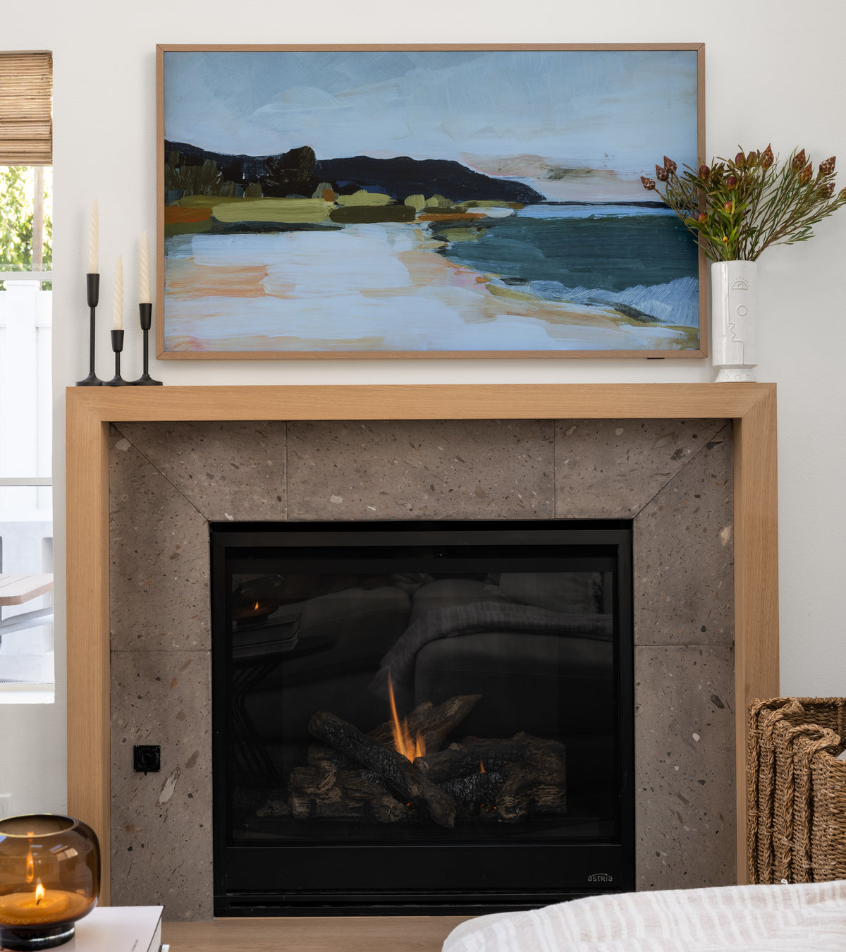A cozy living space features a modern fireplace with a Zia Tile Badlands 12x24 gray stone tile surround and wooden mantel, topped with a coastal painting, candles, and flowers. Nearby sit a wicker basket and glass container.