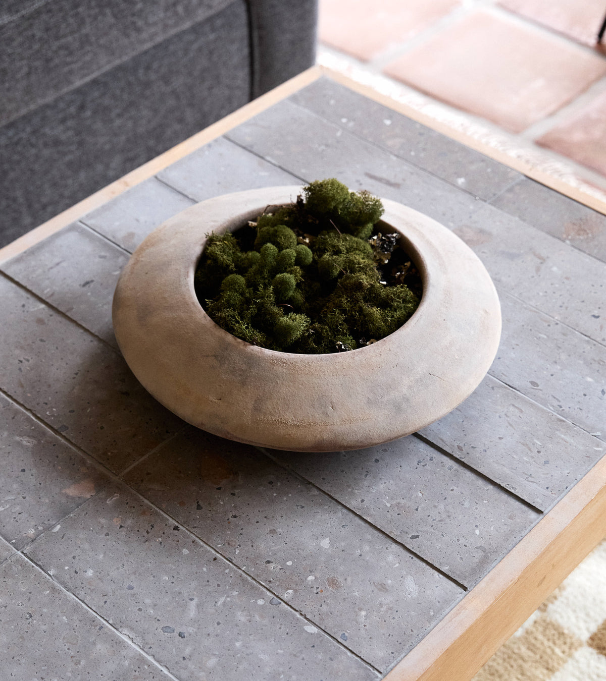 A Zia Tile Badlands 4x12 planter with green moss sits on a gray tiled coffee table beside a dark fabric sofa.