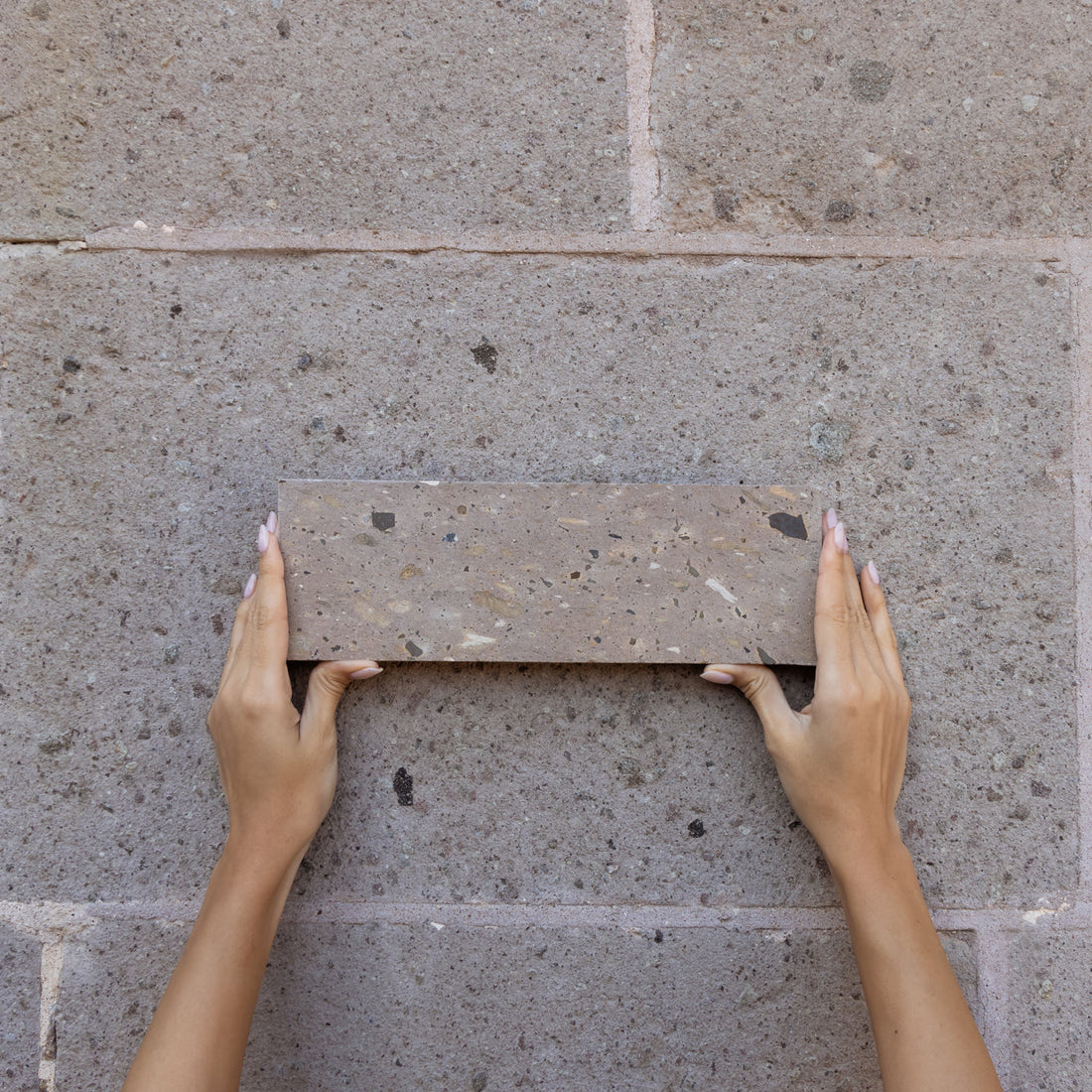 Two hands with manicured nails hold a rectangular brick against a stone wall, matching the color and texture of the background.