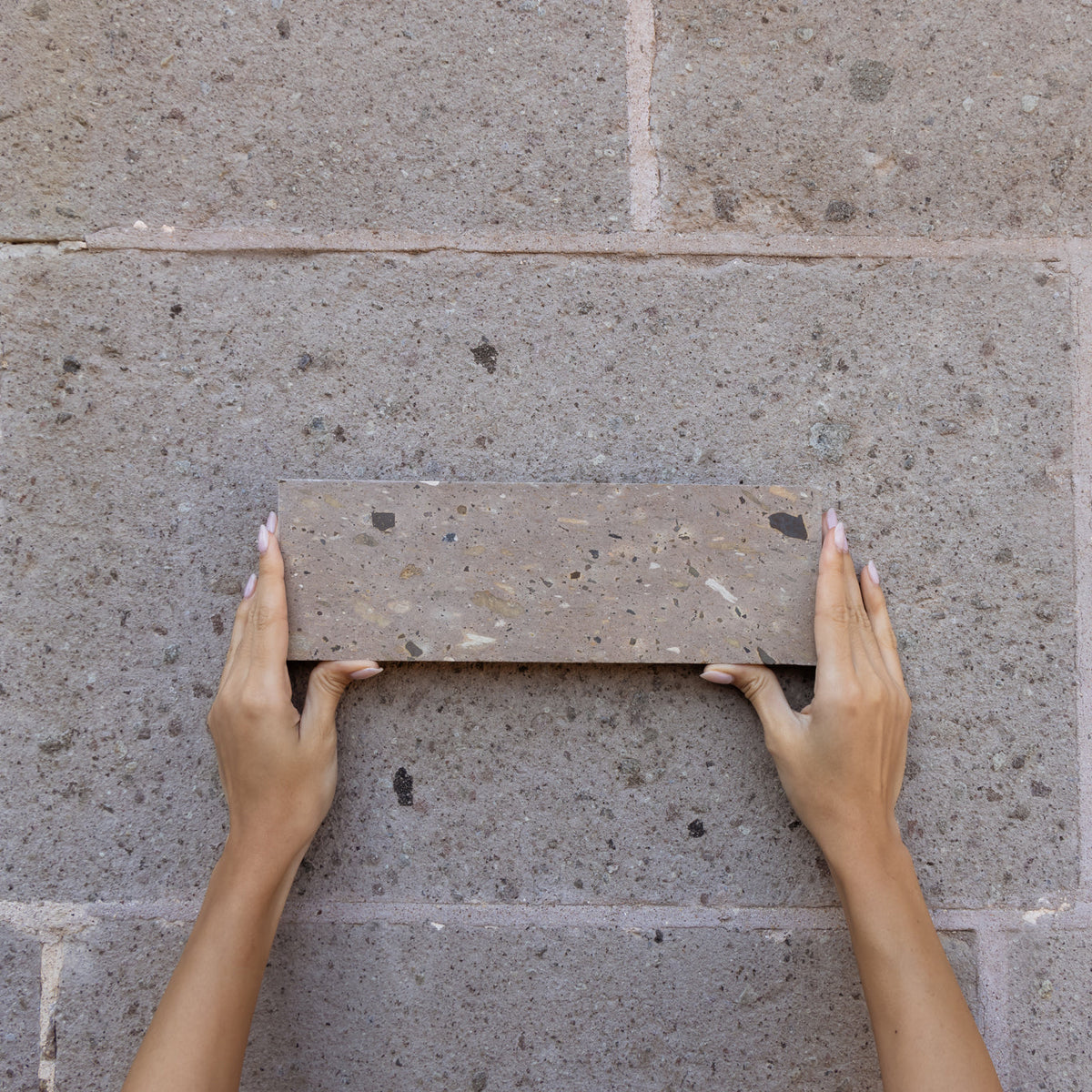 Two hands with manicured nails hold a rectangular brick against a stone wall, matching the color and texture of the background.