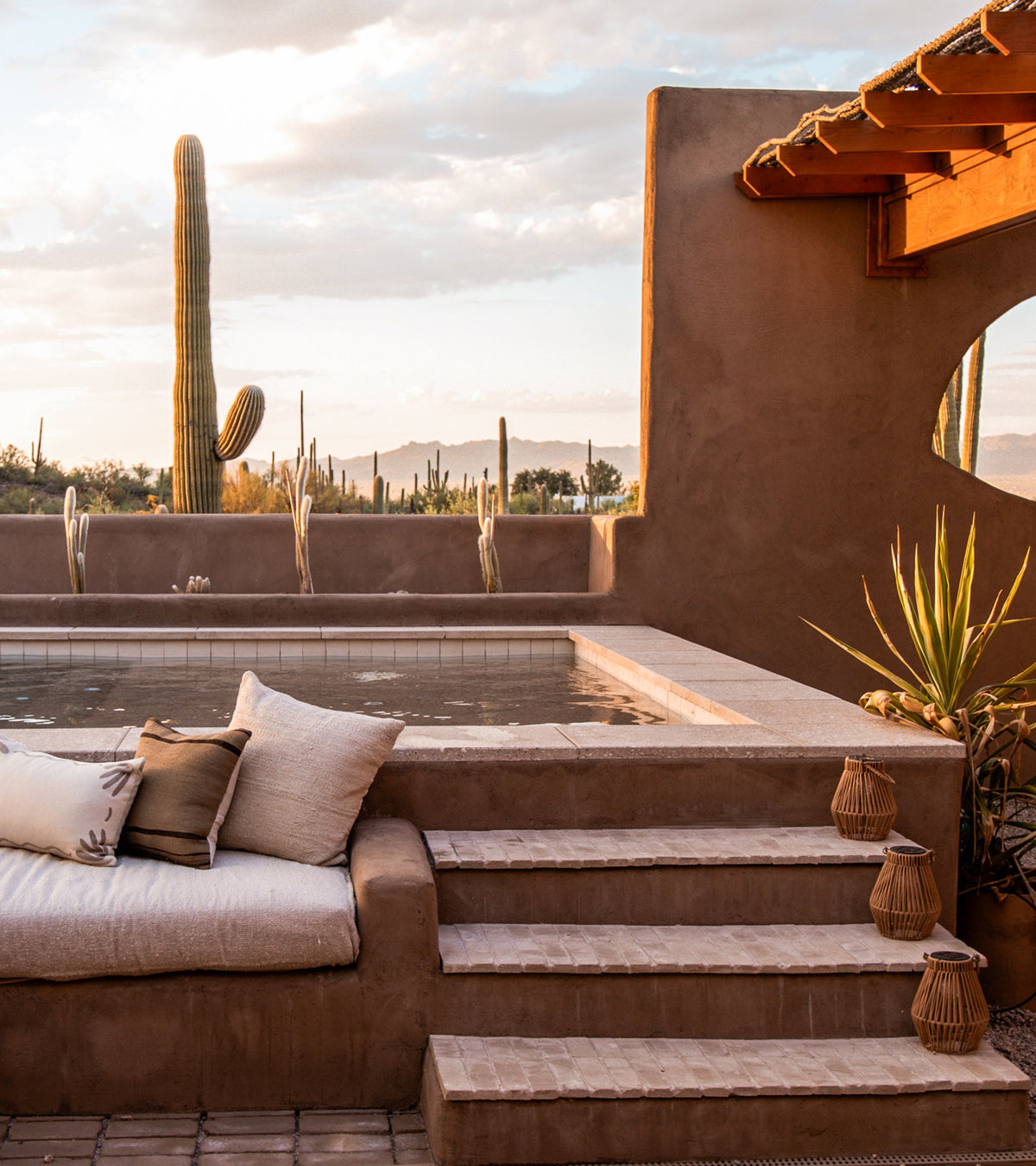 A desert patio at sunset features Zia Tile’s Baja 4x4 Square handmade cotto terracotta steps, a small pool, beige-cushioned built-in bench, lanterns, and cacti under soft clouds.