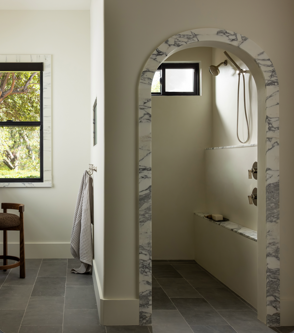 A modern bathroom features Basilica 12x24 + Honed large format stone tiles by Zia Tile on the gray floor, a marble-trimmed arched shower entrance, a towel on the wall, a wooden chair, and two windows letting in natural light.