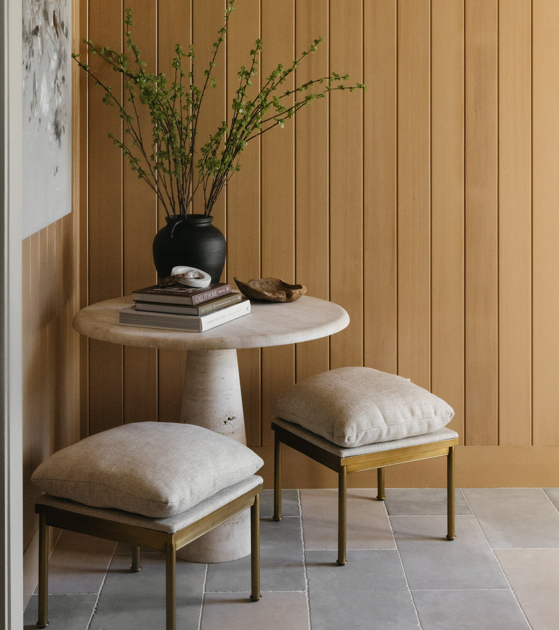 A small round marble table with books, a black vase of green branches, and a wooden bowl sits against wood paneling, flanked by two cushioned stools on Zia Tile’s Basilica 12x24 + Honed tiles for a sleek, modern look.