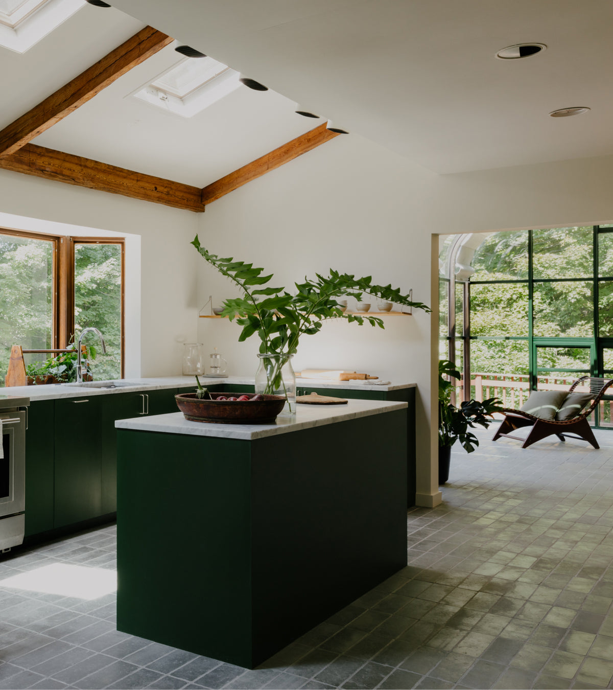 A kitchen island with a white countertop and a green base sits on a floor of Basilica 4x12 + Honed Moroccan limestone tiles in a sunlit kitchen with dark green cabinetry.