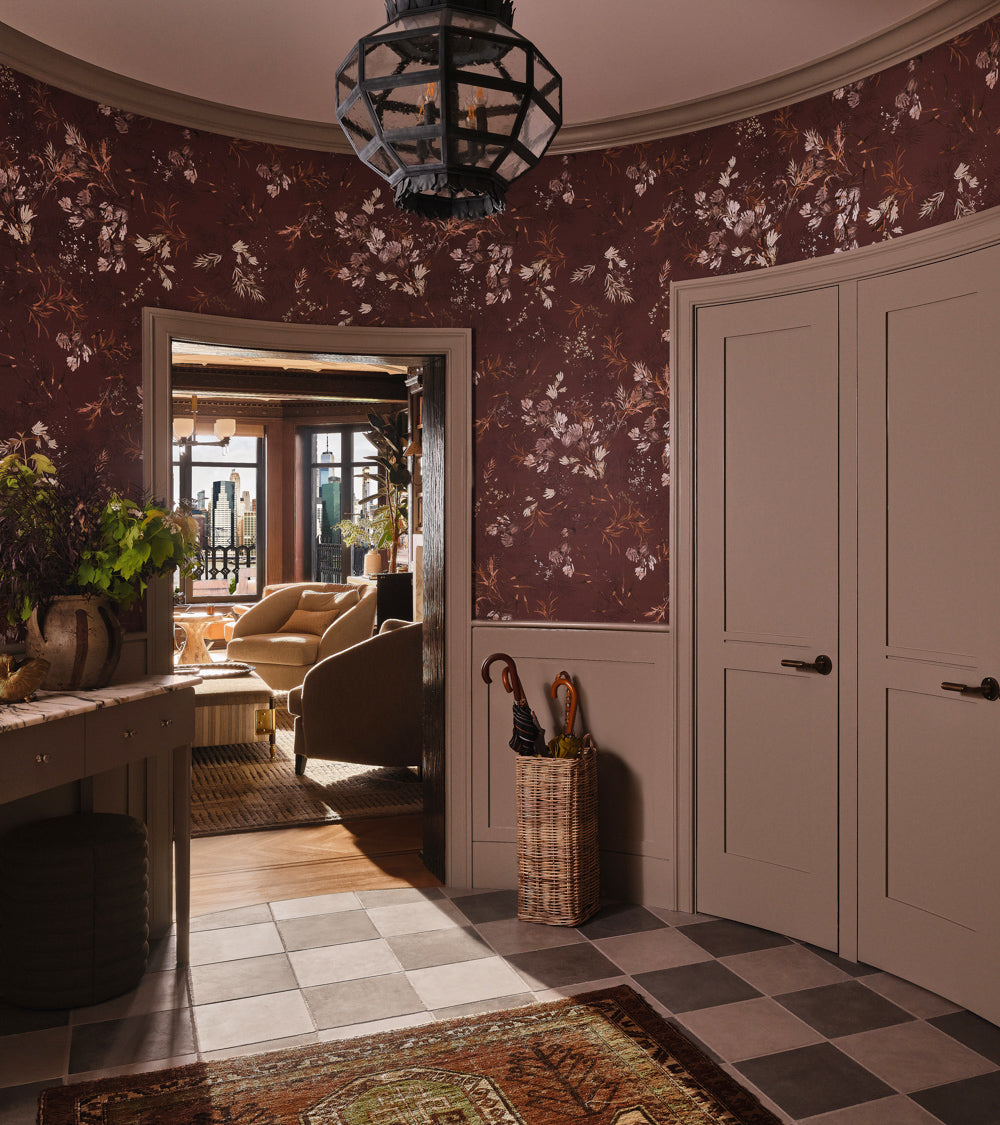 A foyer features floral wallpaper, beige doors, and Zia Tile’s Basilica 12x12 + Honed floors. A wicker umbrella stand sits nearby. Light pours in from the living room with tan sofas and large windows.