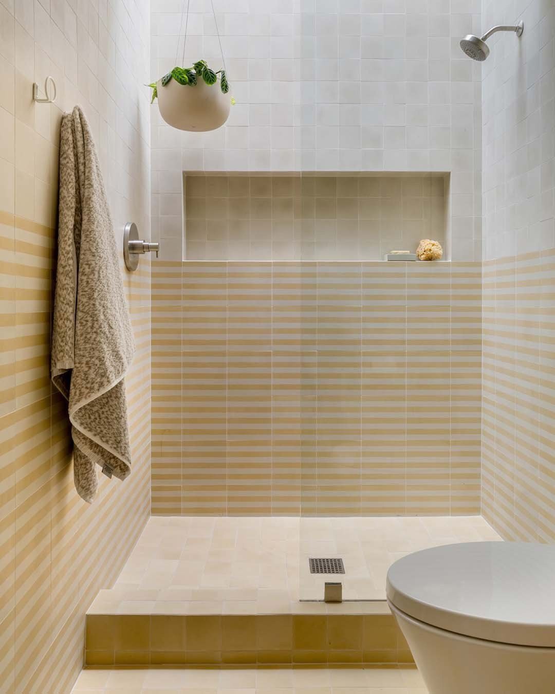 The clean, minimal shower features Zia Tile’s Bateau Mojave 8x8 beige striped geometric tile, a built-in niche, wall-mounted showerhead, gray towel on a hook, hanging potted plant, and partial view of a white toilet.