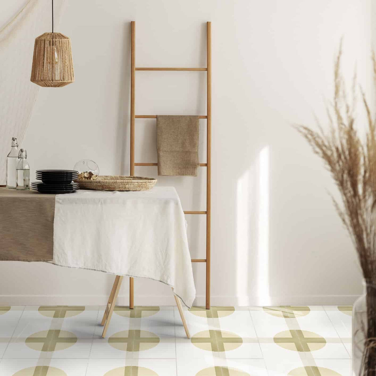 Minimalist dining area featuring a table with a beige and white cloth, black dishes, a woven basket, wooden ladder with towel, rattan pendant lamp, dried grass in sunlight, and a geometric floor with Bauhaus White 8x8 tiles by vendor-unknown.