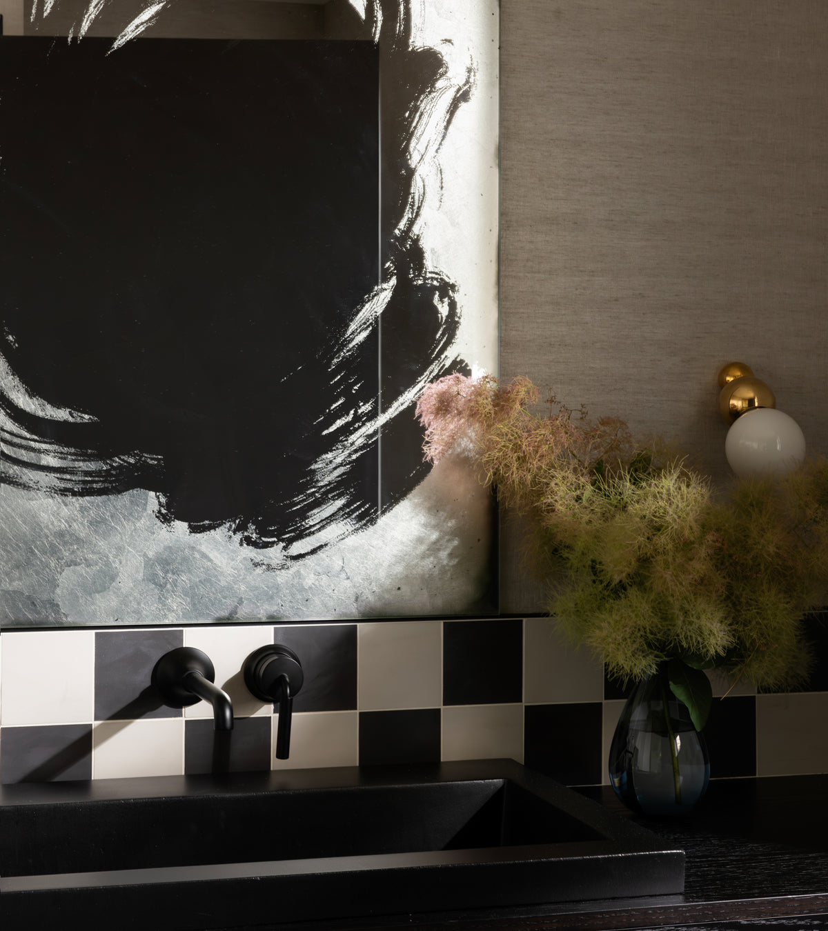 A modern bathroom features a black sink, matte black faucet, vendor-unknown Black 4x4 tile backsplash, an abstract mirror, a gold and white sconce, and a vase with feathery pink and green foliage.