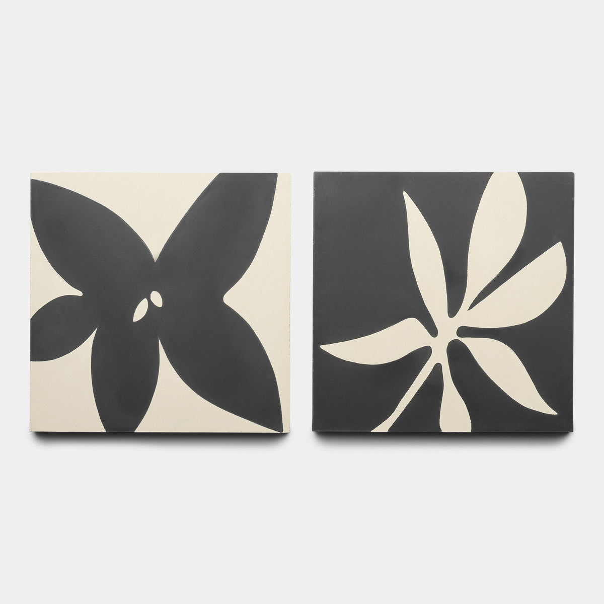 The Flora 8x8 Set Black + Bone, Sample by Zia Tile includes two square tiles with bold black and bone floral designs: one features a five-petal flower, the other a six-petal flower, each on contrasting backgrounds.