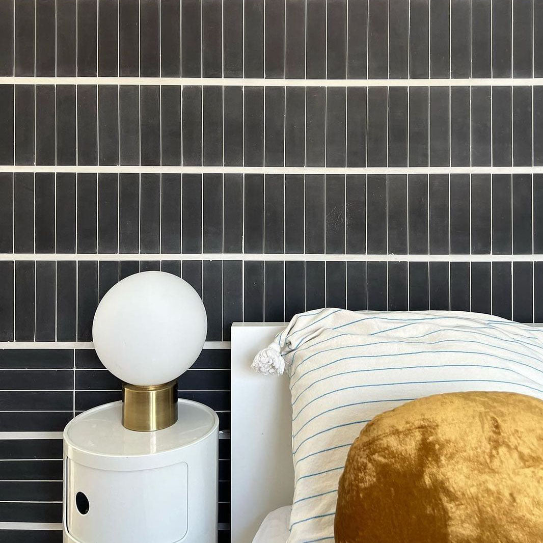 A modern bedroom with a Zia Tile Black 2x8 handmade tile wall, a white bed featuring a blue-striped pillow and round mustard-yellow cushion, plus a white nightstand topped with a spherical brass-based lamp.