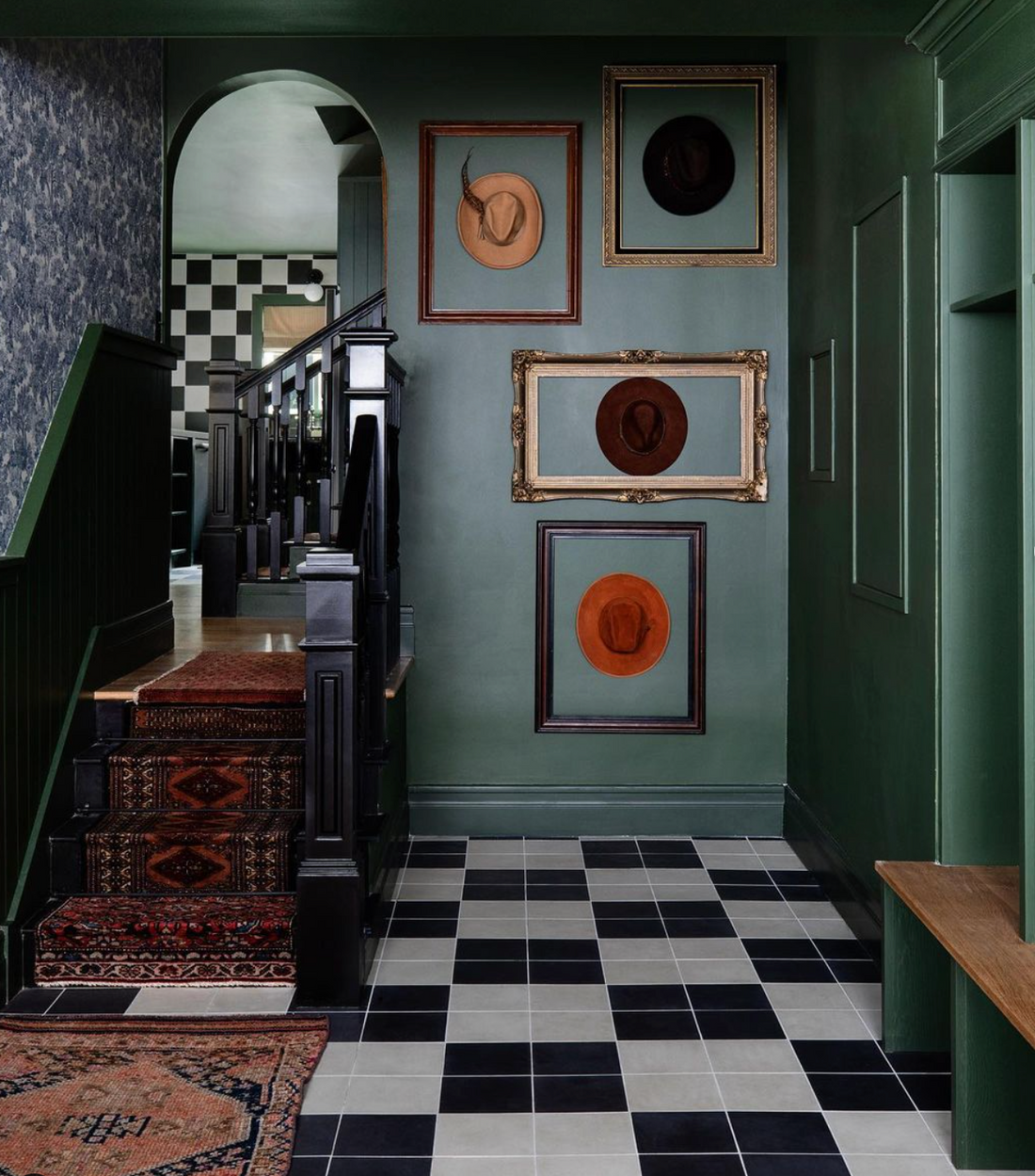 A hallway with black and white checkered tile floor, dark green walls, a staircase with patterned carpets, and framed portraits of hats on the wall. The space features vintage and eclectic decor.